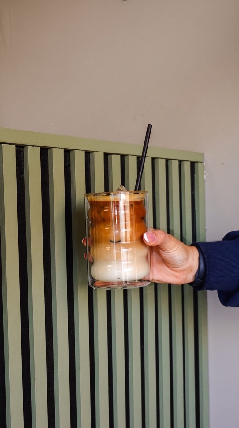 Iced layered coffee held against the sage-green slatted wall, on our Instagram