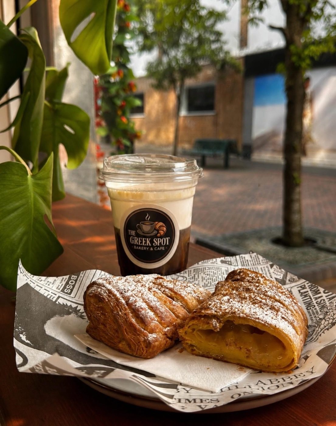 Custard bougatsa dusted with icing sugar and a freddo espresso in a Greek Spot cup, on newspaper-print paper in the window light